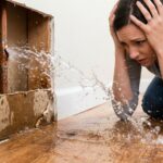 A distressed woman holds her head in panic as water forcefully sprays from a burst copper plumbing pipe inside an open section of a drywall wall, flooding a hardwood floor.