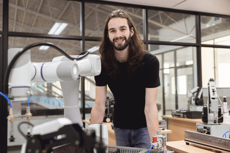 Smiling engineer standing beside a collaborative robotic arm in a modern robotics laboratory, with automation equipment and testing stations visible in the background.