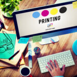 alt=Graphic designer working on a desktop monitor displaying CMYK printing color process with office supplies on a wooden desk