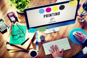 alt=Graphic designer working on a desktop monitor displaying CMYK printing color process with office supplies on a wooden desk