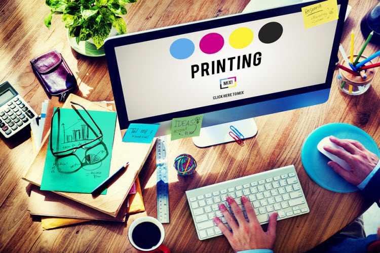 alt=Graphic designer working on a desktop monitor displaying CMYK printing color process with office supplies on a wooden desk