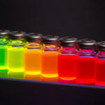 Colorful laboratory vials filled with glowing fluorescent liquids arranged in a row against a dark background.