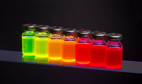 Colorful laboratory vials filled with glowing fluorescent liquids arranged in a row against a dark background.